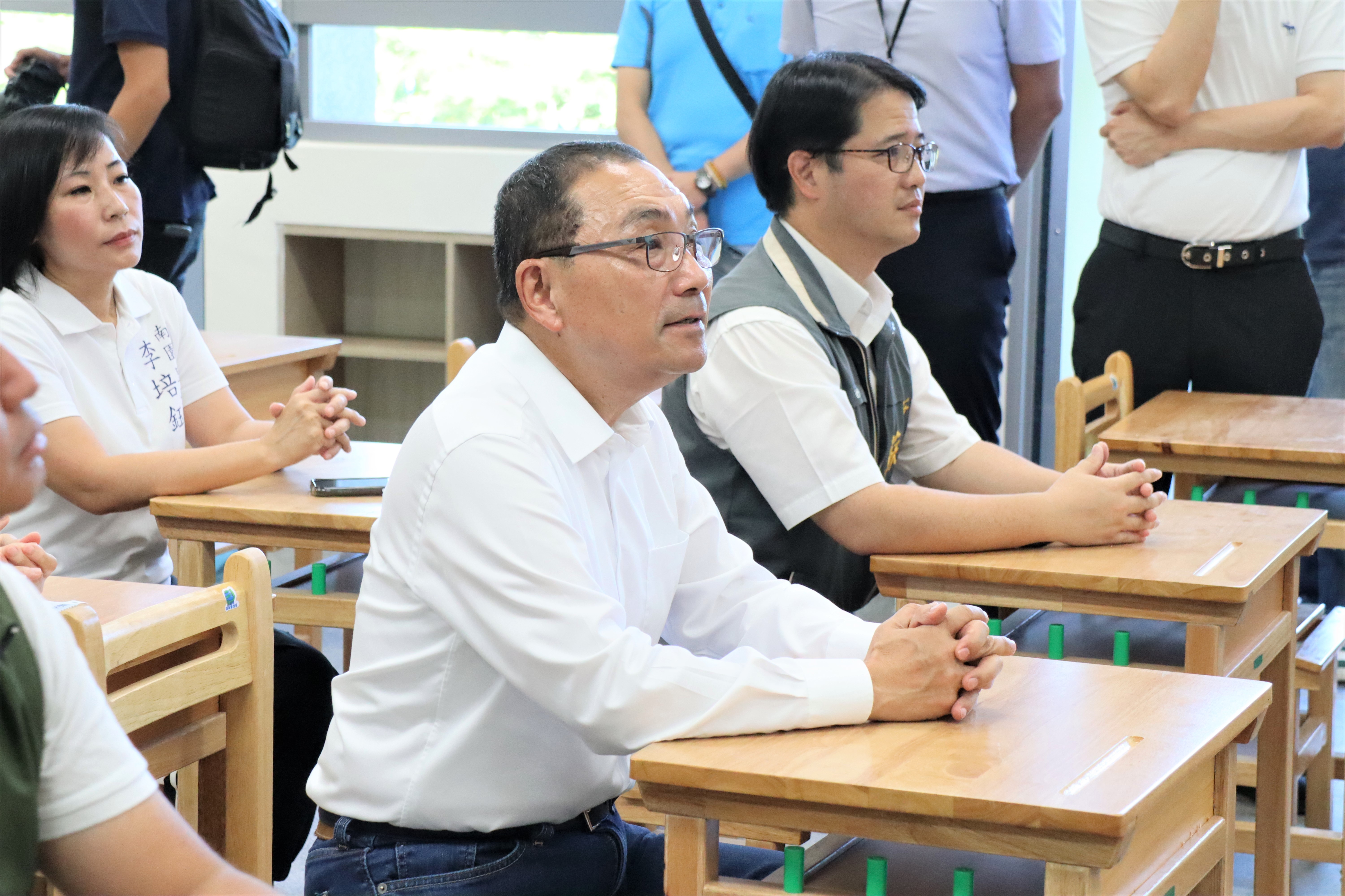 市長侯友宜至北大特區桃子腳國中小視察校舍完工，與多位民代一起坐在新教室中模仿學生上課般，聽取校長簡報。